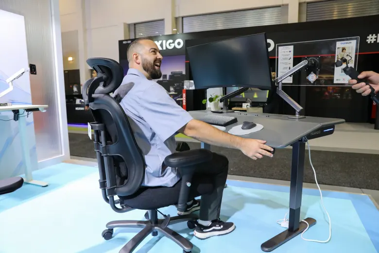 Mężczyzna odchylony na czarnym krześle ergonomicznym HUANUO przy biurku z monitorem na ramieniu, na stoisku IFA 2026