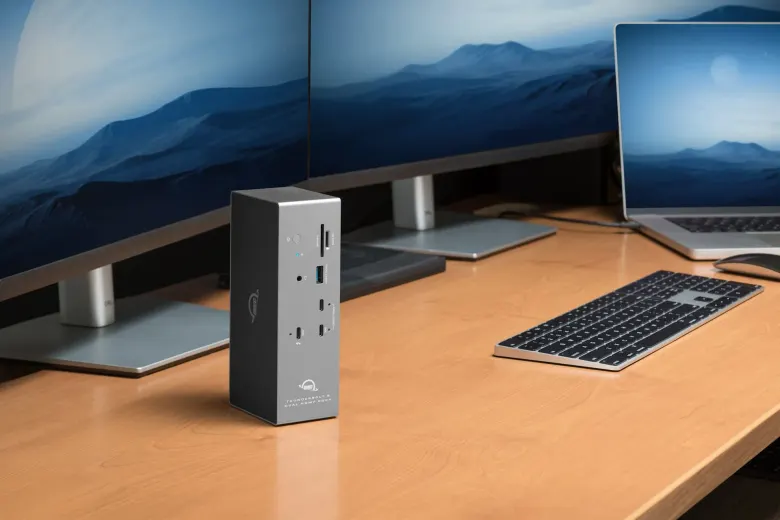 Stacja OWC Thunderbolt 5 Dock postawiona pionowo na drewnianym biurku obok laptopa i dwóch monitorów