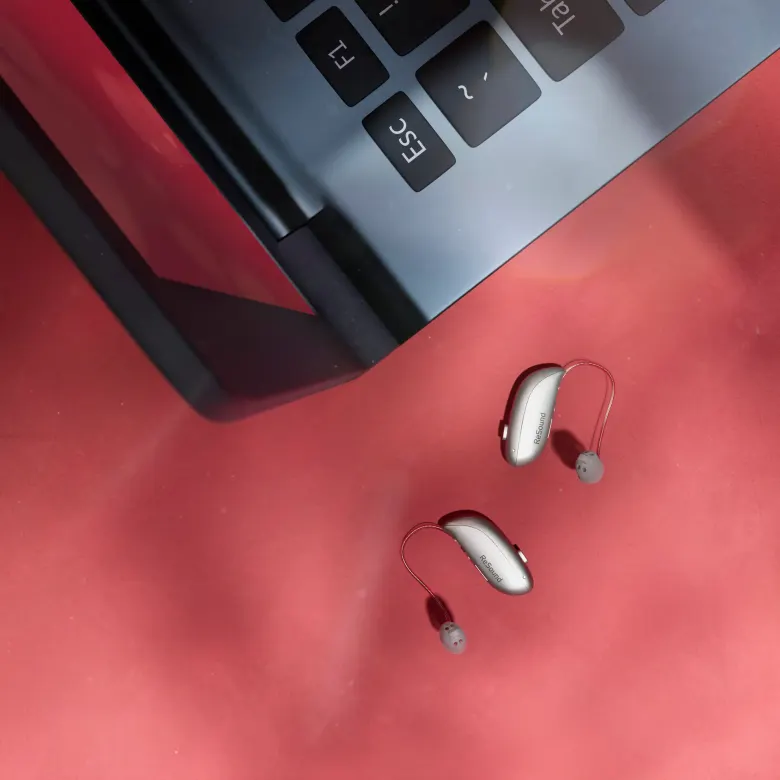 Dwa aparaty słuchowe ReSound Sensia microRIE leżą obok laptopa na czerwonym blacie