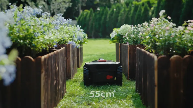 Kosiarka XcanMow Mix 2000 przejeżdża wąskim przejściem między drewnianymi rabatami z kwiatami, napis 55 cm